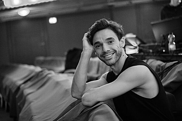 Ein Mann sitzt nach vorn gebeugt auf einem Stuhl im Theater. Eine Hand stützt seinen Kopf. Er schaut lächelnd in die Kamera.