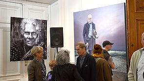 Menschen in einer Ausstellung. Im Hintergrund hängt links eine Fotografie. Und rechts ein Gemälde.