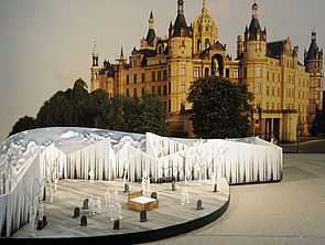 Das Bühnenmodell steht vor einem Foto vom Schweriner Schloss. Zum Modell gehört eine halbkreisförmige, weiße Wand. Auf der Bühne stehen Bäume, Grabsteine, Figuren und ein Doppelbett.