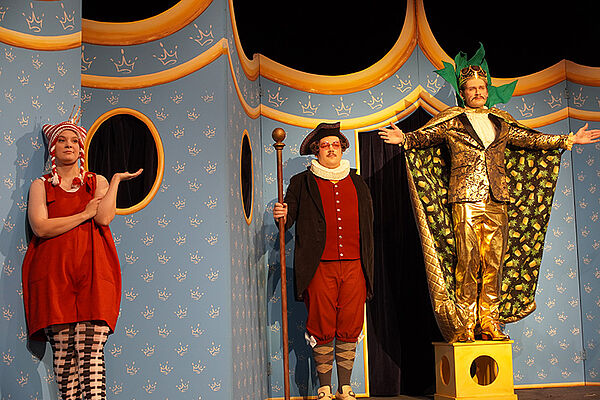 Drei Schauspieler. Sie stehen in Kostümen auf einer Bühne. Sind sind: ein Kaiser, ein Minister, eine Prinzessin.