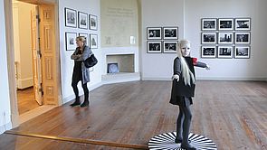 Eine Frau steht in einem Ausstellungsraum und betrachtet Bilder. In der Mitte des Raumes steht eine Figur. 