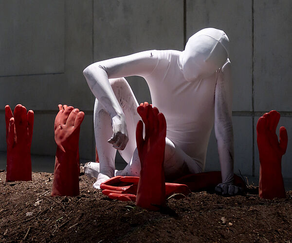 Eine Figur in einem weißen Ganzkörperanzug hockt auf einem Berg Erde, aus dem rote Handschuhe herausstehen.