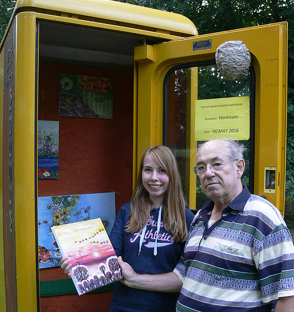 Ein Mann und eine Frau stehen mit einem etwa A4 großen Bild in der Hand an einer gelben, geöffneten Telefonzelle. 