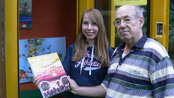 Ein Mann und eine Frau stehen mit einem etwa A4 großen Bild in der Hand an einer gelben, geöffneten Telefonzelle.