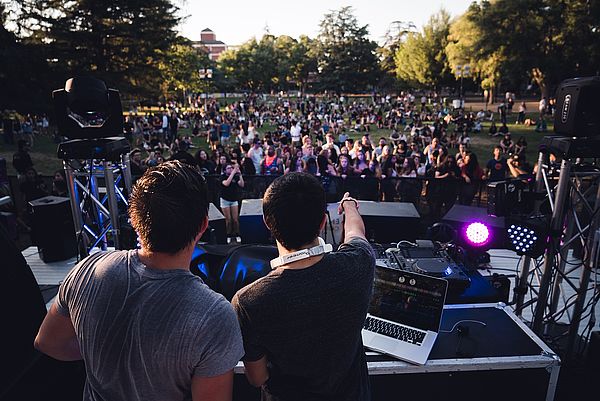 Zwei DJs schauen vom Mischpult aus auf tanzende Menschen in einem Park.