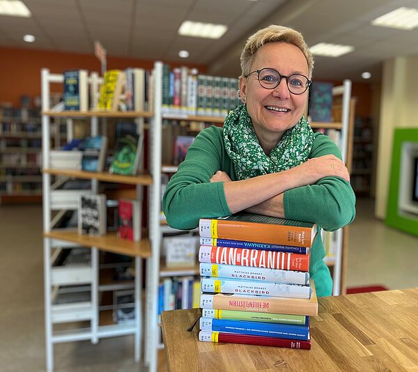 Grit Wilke steht vor einem Tisch und stützt sich auf einen Stapel Bücher. Im Hintergrund stehen Bücherregale.