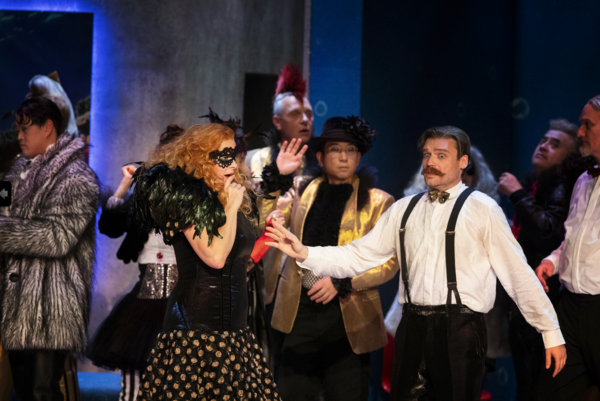 Männer und Frauen auf der Bühne. Manche sind verkleidet. Sie singen und spielen.