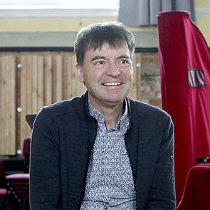 Volker Reinhold sitzt in einem Probenraum. Hinter ihm steht eine abgedeckte Harfe.