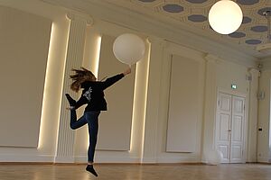 Ein Mädchen tanzt mit einem weißen Luftballon in der Hand durch einen Saal.