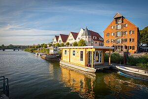 Ein Fluss. An dem Fluss liegen Boote und stehen Häuser.