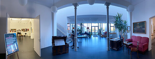 Ein großer Raum mit einem großen Durchgang, der von zwei Säulen getragen wird. Links ist eine Doppeltür geöffnet, dahinter befindet sich eine Ausstellung. Ein rotes Sofa steht im Raum. Treppen führen eine Etage höher. Weiter hinten stehen Tische und Stühle vom Café.  