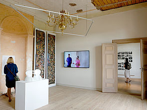 Ein Ausstellungsraum. Im Raum steht ein weißer Sockel mit einer weißen Skulptur. An der Wand hängen ein Bildschirm und Gemälde. Im Raum steht eine Frau.