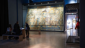 Ein übermannshoher Webteppich aus dem Mittelalter hinter einer Glaswand. Davor Ausstellungsbesucher.