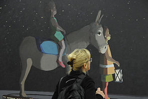 Eine Frau betrachtet ein Gemälde. Darauf sitzt eine Person auf einem Esel, eine Person läuft neben ihm.