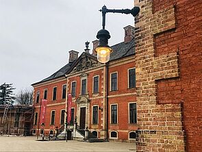 Detailaufnahme einer Laterne an einer Backsteinwand. Im Hintergrund: der Eingang zum Schloss.