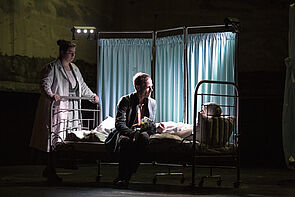 Theaterszene: Ein Mann sitzt auf einem Krankenbett. Eine Frau steht am Fußende.
