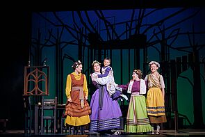 Fünf junge Frauen und Mädchen stehen auf der Bühne. Sie tragen bunte Kleider. Sie sind Schauspielerinnen.