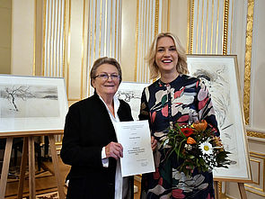 Inge Jastram und Ministerpräsidentin Manuela Schwesig stehen nebeneinander. Inge Jastram hält eine Urkunde in der Hand, Manuela Schwesig einen Blumenstrauß. Hinter ihnen stehen Zeichnungen der Preisträgerin.