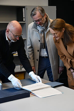 Drei Personen beugen sich über einen Tisch und betrachten historische Dokumente in einer geöffneten Archivmappe. Zwei Personen tragen weiße Baumwollhandschuhe.