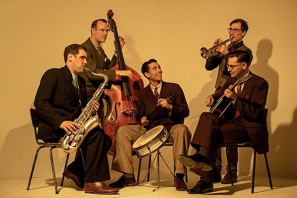 5 Musiker sitzen im Kreis und spielen Instrumente.