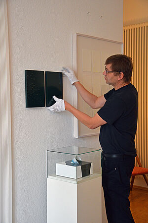 Christian Frosch hat weiße Handschuhe an und greift nach einem kleinen, dunklen Bild an der Wand.