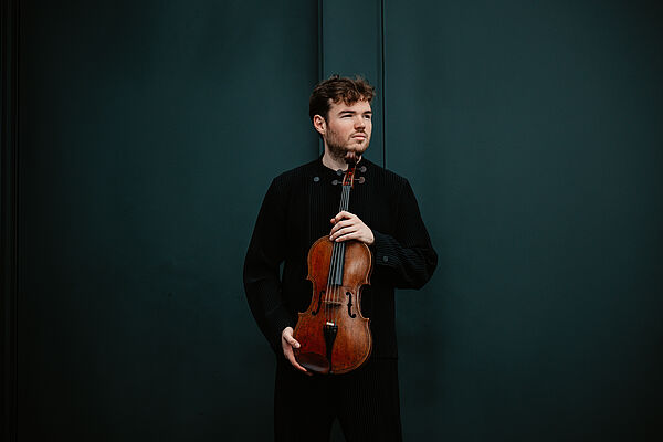 Porträt eines Musikers. Er ist dunkel gekleidet, hält eine Bratsche in seinen Händen.