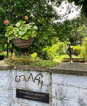 Ein Garten. Davor steht eine halbhohe Mauer. An der Mauer hängen die Initialen des Künstlers: ONH. Und ein Schild mit dem Hinweis auf die Gedenkstätte.