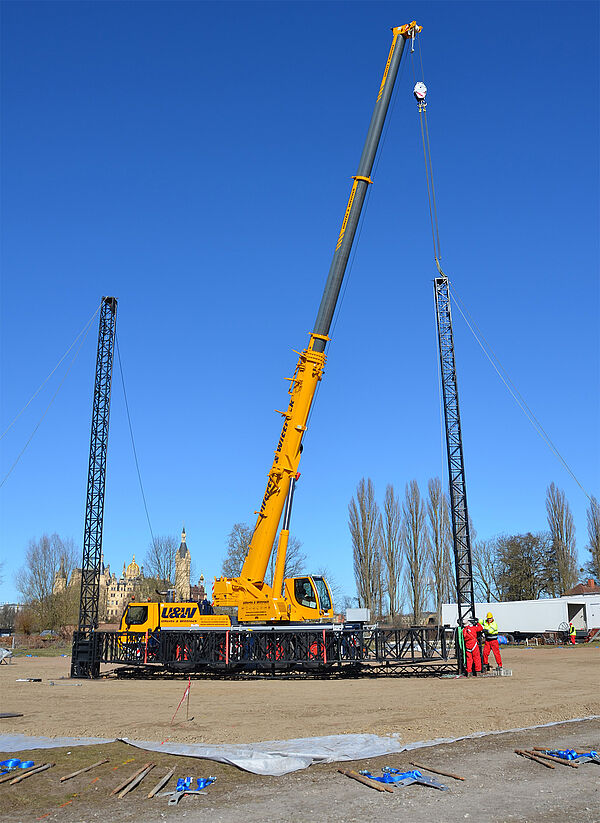 Ein großer Platz. Auf dem Platz steht ein Kran. Der Kran und Bauarbeiter stellen die ersten Pfeiler für das Theaterzelt auf. 