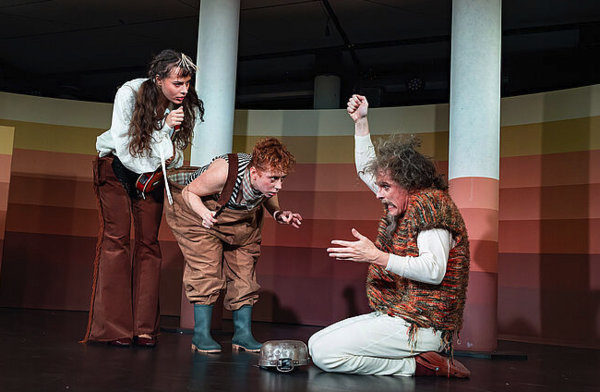 Drei Schauspieler auf der Bühne. Einer spielt, zwei starren ihn an. 