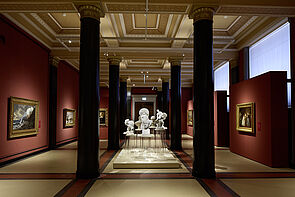 Ein Ausstellungsraum. An den Wänden hängen Ölgemälde. In der Mitte stehen Gipsplastiken.