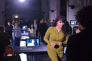 Eine Frau vor der Kamera. Im Hintergrund stehen mehrere Personen.