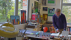 Matthias Kanter blättert in seinem Atelier in Zetteln. An den Wänden hängen Bilder. Auf dem Tisch liegen Pinsel und Farben.