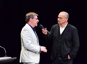 Zwei Männer stehen auf einer Bühne und sprechen miteinander. 