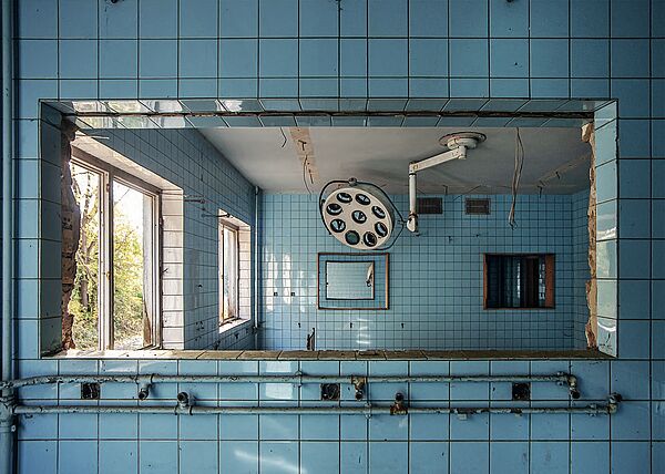 Ein verfallener Operationssaal. An den Wänden befinden sich blaue Fliesen. An der Decke hängt OP-Schwenklicht.
