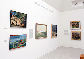Ein Ausstellungsraum. Gemälde hängen an weißen Wänden.