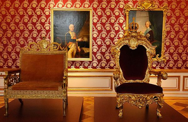 Ein Zimmer mit rot-goldener Tapete. An der Wand hängen zwei Gemälde. Davor stehen auf rotem Teppich zwei goldene Sessel.
