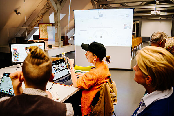 Menschen in einem Raum. Sie sitzen an schreibtischen mit Laptops.