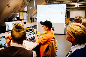 Menschen in einem Raum. Sie sitzen an schreibtischen mit Laptops.