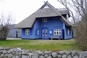 Ein blaues Haus. Das Haus hat weiße Fenster. Das Dach ist aus Reet.