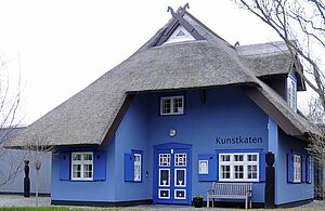 Ein blaues Haus. Das Haus hat weiße Fenster. Das Dach ist aus Reet.