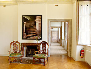 Ein Kamin. Darüber ein Bild mit dem Beginn einer Wendeltreppe. Vor dem Kamin stehen zwei historische Holzstühle mit kunstvollen Holzverzierungen an der Lehne und grünem Polster. Hinter einem geöffneten Türrahmen schließt sich Raum an Raum - ebenfalls von Türrahmen zu Türrahmen. 