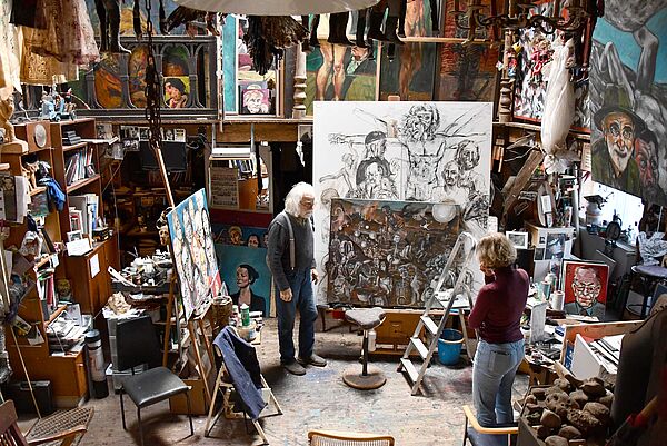 Hans W. Scheibner steht in seinem Atelier. Er spricht mit einer Frau. Rund um sie hängen und stehen viele Gemälde.