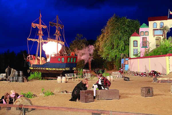 Die Freilichtbühne. Im Hintergrund brennt ein Schiff. Am rechten Bildrand stehen Gebäude. Menschen ducken sich.