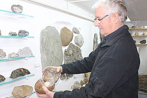 Reinhard Braasch steht vor einer Wand mit großen und kleinen, hellen und dunklen Steinen. In der Hand hält er ein geöffnetes steinernes Ei. 