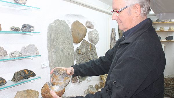 Reinhard Braasch steht vor einer Wand mit großen und kleinen, hellen und dunklen Steinen. In der Hand hält er ein geöffnetes steinernes Ei. 