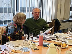Dr. Regina Erbentraut und Dr. Gerhard Graulich schauen auf ein A4-Blatt und unterhalten sich. Vor ihnen auf dem Tisch liegen ein Buch, ein randvoll gefüllter Stehordner und viele Unterlagen.