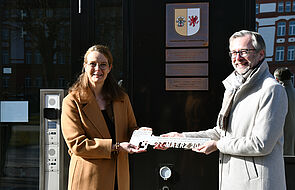 Zwei Personen stehen vor dem Eingang eines Gebäudes mit dem Wappen von Mecklenburg-Vorpommern. Eine Frau und ein Mann halten gemeinsam einen großen Symbolschlüssel mit der Aufschrift „Neubau Depots und Werkstätten, 27. März 2026".