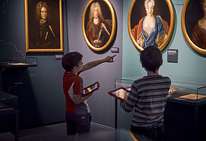 Zwei Kinder stehen mit einem Tablet in der Hand vor einer Bildergalerie adliger Frauen und Männer. 