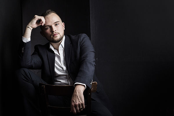 Nils Wanderer sitzt in einem dunklen Anzug vor einem dunklen Hintergrund. Er trägt ein weißes Hemd und stützt lehnt eine Hand an seinen Kopf.
