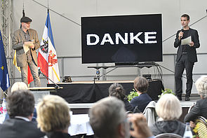 Dr. Marcus Doering und Lars Scheibner stehen zur Preisverleihung auf einer Bühne. Doering links, Scheibner rechts. Zwischen ihnen steht ein schwarzer Bildschirm. Auf ihm steht in weißer Schrift das Wort "Danke". Vor ihnen sitzt Publikum.